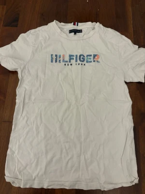 Vit Hilfiger T-shirt med tryck - Vit T-shirt från Tommy Hilfiger med färgglatt tryck på bröstet där det står 'HILFIGER NEW YORK'. Klassisk rund hals och korta ärmar. Perfekt basic-plagg med snygg logga framtill.