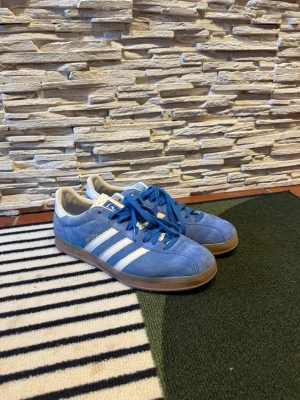 Blå Adidas Gazelle sneakers - Säljer ett par klassiska Adidas Gazelle sneakers i blå mocka med vita detaljer och gummisula. Skorna har de ikoniska tre ränderna på sidan och Adidas-logga på hälen. Perfekta för dig som gillar retrostil och streetwear.