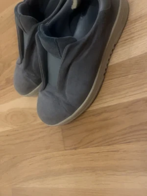 Grå slip-on sneakers från Axel Arigato - Snygga grå slip-on sneakers från Axel Arigato i mocka med elastisk panel över vristen. Skorna har en minimalistisk design och en ljusgrå sula som ger en clean look. Perfekta för dig som gillar stilrena och bekväma sneakers.