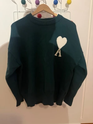 Grön stickad tröja från Ami Paris - Mörkgrön stickad tröja från Ami Paris med vit hjärtlogga och bokstaven A på bröstet. Tröjan har rund halsringning och ribbade muddar. Perfekt för dig som gillar stilrena och trendiga plagg.