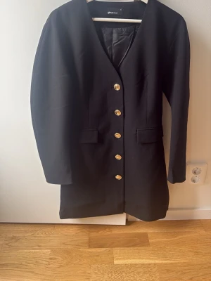 Svart klänning med guldknappar - Stilren svart klänning från Gina Tricot med v-ringning och långa ärmar. Klänningen har dekorativa guldknappar framtill och två fickdetaljer. Passar perfekt för dig som gillar en klassisk look med en twist.