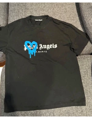 Svart Palm Angels t-shirt med blått hjärta - Cool svart t-shirt från Palm Angels med tryck i vitt och ett blått, droppande hjärta på bröstet. Klassisk passform och rund halsringning. Perfekt för dig som gillar streetwear och vill sticka ut med en unik detalj.