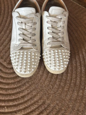 Vita sneakers med nitar från Louboutin - Unika vita sneakers från Christian Louboutin med coola vita nitar på tån och klassisk röd sula. Skorna har lågt skaft, vita skosnören och är tillverkade i läder. Perfekta för dig som vill sticka ut med en edgy och lyxig stil. Man får med sko lådan