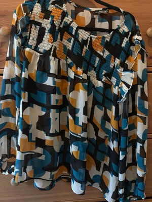 Mönstrad blus från Norah - Cool blus från Norah med grafiskt mönster i blått, svart, vitt och senapsgult. Blusen har smock upptill och volangdetaljer vid axlarna. Långärmad och luftig modell, perfekt för dig som gillar färg och mönster.