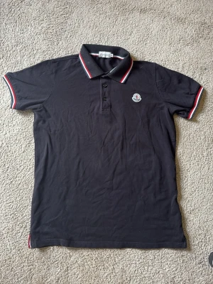 Svart Moncler pikétröja - Snygg svart pikétröja från Moncler med klassisk krage och knappar framtill. Tröjan har vita och röda ränder på krage och ärmslut samt Moncler-logga på bröstet. Perfekt till jeans eller shorts för en clean look.