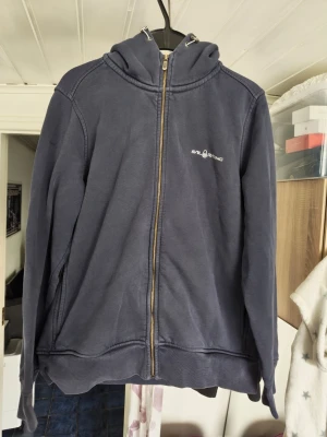 Sail Racing Mörkblå hoodie  - Sail Racing Mörkblå hoodie med dragkedja och huva. Tröjan har två fickor på sidorna och logotyp på bröstet. Materialet är mjukt och bekvämt, perfekt för chill dagar. Klassisk passform och snygga sömdetaljer vid axlarna.
