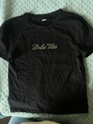 Svart Dolce Vita t-shirt från BikBok - Svart t-shirt från BikBok med texten 'Dolce Vita' i glittriga stenar på bröstet. Klassisk rund halsringning och korta ärmar. Endast använd 1 gång. Den är i storlek M men sitter som S!