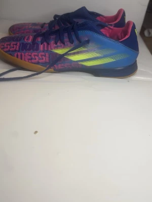 Adidas Messi blå fotbollsskor - Säljer ett par färgstarka Adidas Messi fotbollsskor för inomhusspel. Skorna är blå med neon-gula detaljer och rosa text med Messi-tryck över hela ovandelen. De har blå snörning, rosa innerfoder och en klassisk gummisula för bra grepp på inomhusplaner. Perfekta för dig som vill sticka ut på planen!