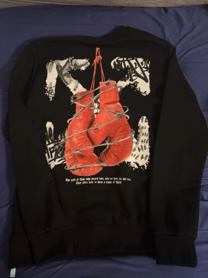 Svart Off-White sweatshirt med tryck - Svart sweatshirt från Off-White med ikoniskt piltryck på bröstet och stort grafiskt motiv med boxningshandskar och taggar på ryggen. Rund halsringning och ribbade muddar. Perfekt för dig som gillar streetwear och unika prints.