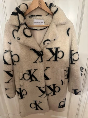 Beige teddyjacka från Calvin Klein - Supermjuk teddyjacka i beige med stora svarta CK-loggor över hela plagget. Jackan har krage, dolda knappar och två fickor framtill. Perfekt för dig som vill ha en trendig och cozy look med streetkänsla.
