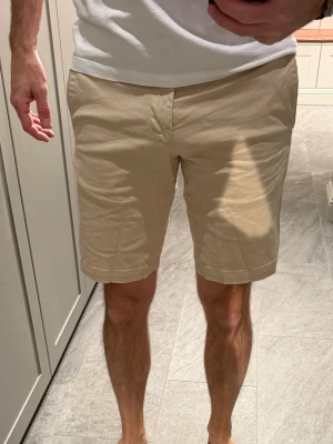 Snygga beige bomullsshorts med fickor - Snygga beige shorts i bomull. Shortsen har sidofickor, bälteshällor och stängs med knapp och dragkedja. Perfekta för varma dagar och enkel att matcha med olika stilar.