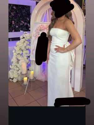Vit axelbandslös långklänning - Elegant vit axelbandslös långklänning med pärldetaljer längs överkanten och en stor dekorativ rosett baktill. Klänningen har en figurnära passform och är tillverkad i ett slätt material som ger en lyxig känsla.