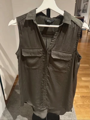 Khakigrön ärmlös blus från H&M - Stilren khakigrön ärmlös blus från H&M med klassisk krage och två bröstfickor. Blusen har en rak passform och knäppning framtill, perfekt för en avslappnad men ändå snygg look.