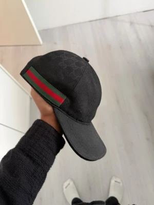 Svart keps från Gucci med GG-mönster - ( BARA KÖPES INGEN BYTE!) Säljer en svart keps från Gucci med diskret GG-monogram över hela tyget. På sidan finns en klassisk grön och röd rand. Kepsen har böjd skärm och justerbar rem bak. Materialet känns som bomullscanvas och designen är riktigt clean och stilren.
