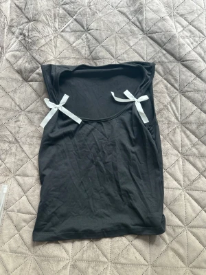 Svart ärmlös topp med rosetter - Snygg svart ärmlös topp med två vita rosetter på axlarna. Toppen har en enkel och stilren design, perfekt att matcha med jeans eller kjol. Materialet känns mjukt och bekvämt mot huden.