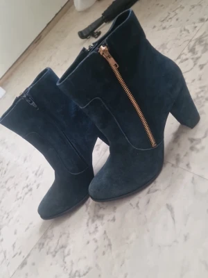 Mörkblåa stövletter med klack - Snygga mörkblå stövletter i mockaimitation med grov klack och guldfärgad dragkedja på sidan. Skorna har rundad tå och en stilren design som passar till många outfits. Perfekta för dig som vill ha en trendig och bekväm look. Mörkgrön färg, nästan svart. Använda ett fåtal gånger. Priset kan diskuteras🫶🏼