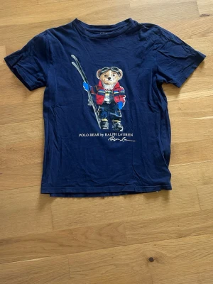 Blå Polo Bear t-shirt från Ralph Lauren - Mörkblå t-shirt från Ralph Lauren med ikoniska Polo Bear-trycket framtill. Klassisk rund hals och korta ärmar. Tillverkad i mjuk bomull som känns skön mot huden. Perfekt för dig som gillar streetstyle och vill sticka ut med en cool printad tee.
