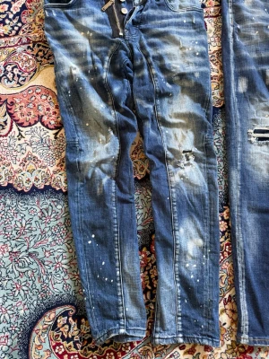 Dsquared2 blå distressed jeans - Säljer ett par blå jeans från Dsquared2 med coolt distressed utseende, slitningar och färgstänk. Jeansen har dragkedja och knappar framtill samt klassisk femficksmodell. Materialet är denim i bomull och passformen är smal.
