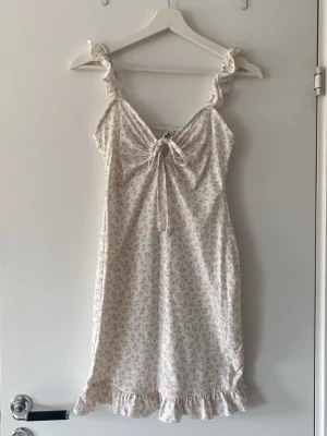 Vit blommig sommarklänning med volang - Söt vit klänning med små rosa blommor, volangdetaljer vid axelband och nederkant samt knyt framtill. Ärmlös modell i lätt material, perfekt för varma dagar. Från Nelly.com i storlek S.