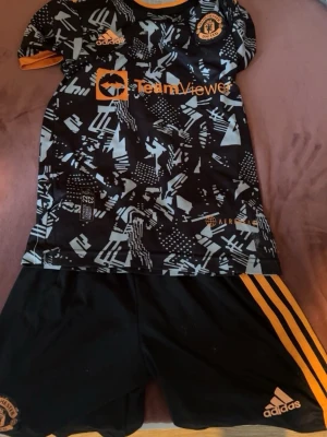 Manchester United träningsset Adidas  - Säljer ett Manchester United träningsset från Adidas med svart och orange mönstrad tröja och svarta shorts med orange detaljer. Tröjan har tryck med TeamViewer och Ronaldo 7 på ryggen. Perfekt för fotbollsträning eller match!