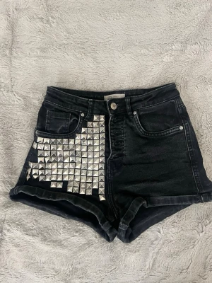 Svarta shorts med nitar - Coola svarta denimshorts med hög midja och uppvikta ben. Ena sidan framtill är dekorerad med silvriga fyrkantiga nitar för en edgy look. Perfekta för dig som vill sticka ut och addera lite attityd till outfiten.