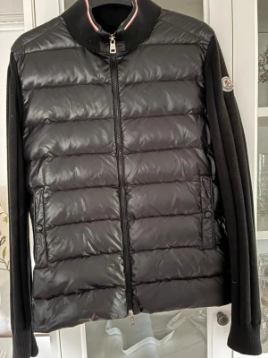 Svart moncler cardigan  - Snygg svart dunväst från Moncler med quiltad design och dragkedja framtill. Västen har två fickor med knappstängning och Moncler-logga på ärmen. Perfekt för lager-på-lager och streetstyle. Materialet är glansigt och ger en exklusiv känsla.