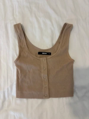 Beige ribbad crop top - Snygg beige ribbad crop top från Bik Bok med knappar framtill. Toppen har breda axelband och rundad halsringning, perfekt för en trendig och avslappnad stil. Materialet är stretchigt och bekvämt.