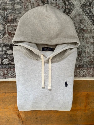 Ljusgrå Polo Ralph Lauren Hoodie - Storlek M - Hej! 👋 Säljer en klassisk ljusgrå Polo Ralph Lauren hoodie i storlek M med marinblå pony-logga. 🐎✨ Tröjan är i mycket gott skick och är välvårdad. Liten slitning på ena ärmslutet, men har knappt märkt det själv under användning. Garanterat 100% äkta med originaltvättlappar kvar (ingen QR-kod på just denna årsmodell). Det absolut mest mångsidiga och stilrena basplagget man kan ha i garderoben – passar till allt! Fraktar snabbt! 💬 100% 🚛📦 