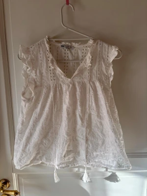 Vit broderad blus med volangärm - Söt vit blus från Only med broderade detaljer och volang vid ärmslut. Blusen har v-ringning med knytband och är tillverkad i ett lätt, luftigt tyg. 