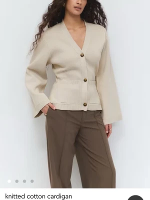 Beige stickad kofta från Gina Tricot - Snygg beige stickad kofta i bomull från Gina Tricot med v-ringning och stora bruna knappar. Koftan har två framfickor och ribbade detaljer vid ärmslut och nederkant. Perfekt för lager-på-lager och enkel att matcha.