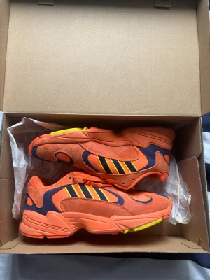 Adidas orange sneakers med gula detaljer - Färgstarka sneakers från Adidas i orange mocka och textil med mörkblå och gula detaljer. Klassisk modell med lågt skaft och ikoniska tre ränder på sidan. Sulan är orange med inslag av gult. Perfekta för dig som vill sticka ut med din stil.