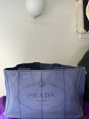 Blå canvas handväska från Prada - Snygg blå handväska från Prada i canvas med tryckt logga framtill och klassisk rektangulär form. Väskan har två handtag och guldiga detaljer upptill. Perfekt för dig som gillar stilrena och trendiga accessoarer.