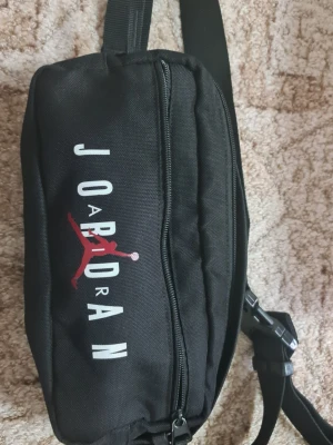 Svart Jordan magväska med logga - Snygg svart magväska från Jordan med vit och röd logga framtill. Väskan har justerbart band med spänne och dragkedja upptill. Perfekt för att bära det viktigaste och har en sportig streetstyle-vibe.