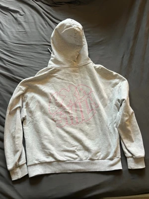 Ljusgrå hoodie med rosa tryck från LXA STHLM (Linn Ahlborg) - Ljusgrå hoodie från märket LXA The Label (Linn Ahlborgs klädmärke). På ryggen finns ett stort rosa grafiskt tryck med texten 'LXA STHLM'. Hoodien har ribbade muddar vid ärmslut och nederkant. 