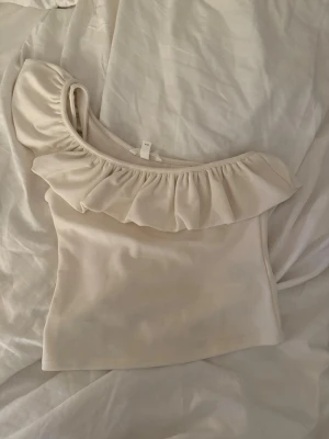 Beige oneshoulder topp med volang - Superfin beige topp från H&M med oneshoulder-design och bred volang längs halsringningen. Sitter tight och är perfekt till sommaren. Toppen är i bra skick o säljs då den nog inte kommer komma till användning mer🤍🤍