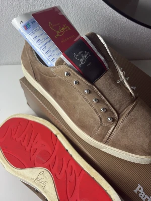 Beige sneakers från Christian Louboutin - Snygga beige sneakers från Christian Louboutin i mocka med ikoniska röda sulor och silverfärgade nitar på sidan. Skorna har en klassisk låg profil och vita kanter runt sulan. Perfekt för dig som vill sticka ut med exklusiva detaljer.