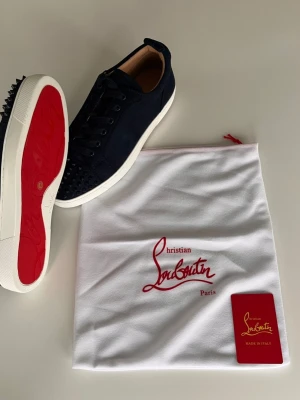 Mörkblå Christian Louboutin sneakers - Snygga mörkblå sneakers från Christian Louboutin med klassiska röda sulor och svarta nitar på tån. Skorna har en låg profil, vita kanter på sulan och är tillverkade i mocka. Perfekta för dig som vill sticka ut med exklusiv design.