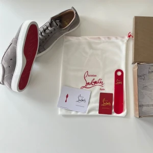 Gråa sneakers från Christian Louboutin - Snygga gråa sneakers från Christian Louboutin med klassisk röd sula. Skorna har en låg profil, grå mocka och vita gummisulor. Perfekta för dig som vill ha en stilren och exklusiv look. Levereras med dustbag och originaltillbehör.