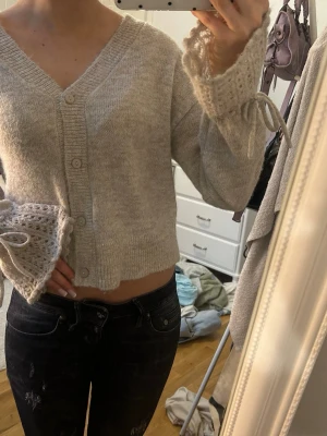 Beige kofta med virkade ärmslut - Mysig beige kofta med v-ringning och knäppning framtill. Koftan har unika virkade ärmslut med rosettband som ger en söt och trendig touch. 