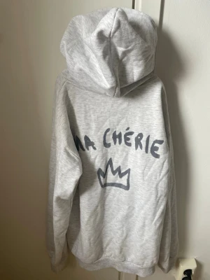 Grå hoodie med tryck 'Ma Chérie' - Ljusgrå hoodie med huva och snörning, tryck med texten 'Ma Chérie' och en krona både fram och bak. Skriv om ni har frågor! storlek 158/164. Den är knappast använd!🤍🤍