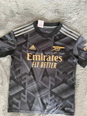 Arsenal svart matchtröja Adidas - Snygg svart Arsenal matchtröja från Adidas med guldiga detaljer och loggor. Tröjan har geometriskt mönster, korta ärmar och rund hals. Materialet är lätt och andas, perfekt för träning eller match. Officiell Emirates sponsorlogga på bröstet.