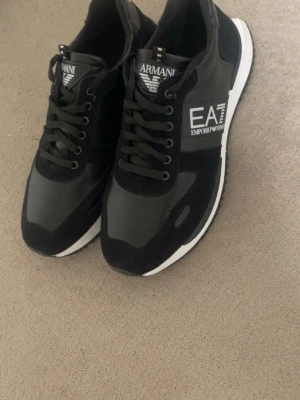 Svarta Armani EA7 sneakers - Snygga svarta sneakers från Armani EA7 med vita detaljer och logga på sidan. Skorna har en klassisk låg modell, svarta skosnören och en vit sula. Materialmix av textil och syntet ger en modern look som passar till många outfits. Helt nya