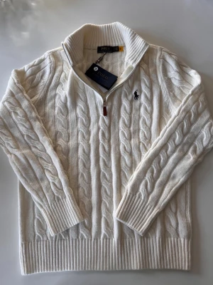 Vit kabelstickad tröja Polo Ralph Lauren - Kabelstickad vit tröja från Polo Ralph Lauren med halv dragkedja och hög krage. Tröjan har ribbade muddar och nederkant samt den klassiska broderade loggan på bröstet. Tillverkad i mjukt material som känns lyxigt och stilrent.