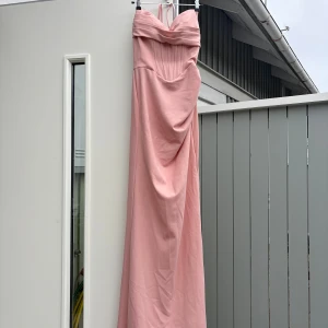 Lång ljusrosa klänning med drapering - Elegant långklänning i ljusrosa med draperad detalj framtill och axelbandslös design. Klänningen har figurnära passform och markerad midja, perfekt för speciella tillfällen. Tillverkad i ett mjukt, följsamt material som ger en lyxig känsla.