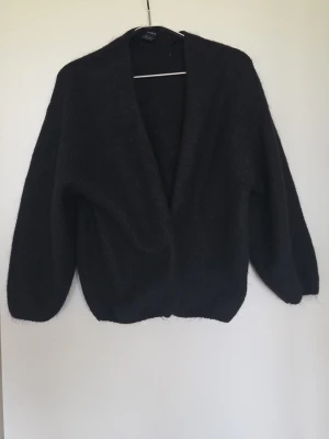Lindex VANESSA cardigan i ull - Elegant svart öppen cardigan från Lindex i storlek S/XS. Koftan har en enkel och stilren design utan knappar, med mjukt och fluffigt material som känns skönt mot huden.