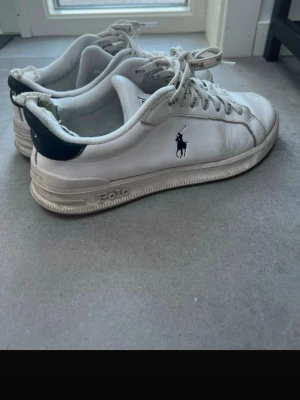 Vita Polo Ralph Lauren sneakers - Snygga vita sneakers från Polo Ralph Lauren med klassisk låg profil. Skorna har svarta detaljer på hälen och Polo-logga på sidan. Tillverkade i läder med vita snören där Polo står tryckt. Perfekta för en stilren och sportig look.