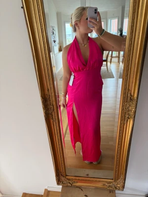 Rosa långklänning med slits - Snygg rosa långklänning med halterneck och djup v-ringning. Klänningen har en hög slits på ena sidan och knyts i nacken. Perfekt för dig som vill sticka ut med en färgstark look. Materialet känns lätt och följsamt.