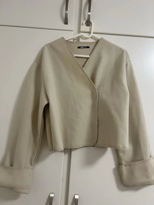 Beige omlottkofta från Gina Tricot - Stilren beige omlottkofta från Gina Tricot med vida ärmar och dekorativa sömmar längs kanterna. Koftan har en avslappnad passform och stängs med dolda tryckknappar framtill. Perfekt för lager-på-lager och enkel att matcha.