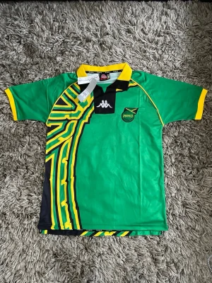 Jamaica fotbollströja från Kappa - Snygg grön Jamaica fotbollströja från Kappa med svart och gul grafisk design på sidan, broderad logga och gul krage. Perfekt för dig som vill sticka ut på planen eller visa ditt stöd för laget. Materialet är lätt och andas, med klassisk sportig passform.