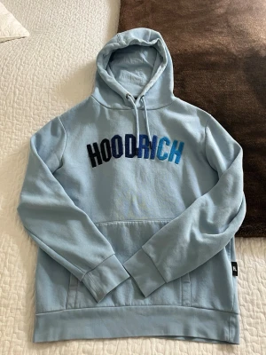Ljusblå hoodie från Hoodrich - Säljer en ljusblå hoodie från Hoodrich med broderad logga i blå toner på bröstet. Hoodien har huva med dragsko, magficka och ribbade muddar. Tillverkad i mjuk bomullsblandning som känns skön mot huden. Den har två små röda fläckar, en på armen och den andra på sidan av magen. Utöver det så är den i fint skick, hör av er om ni har frågor!